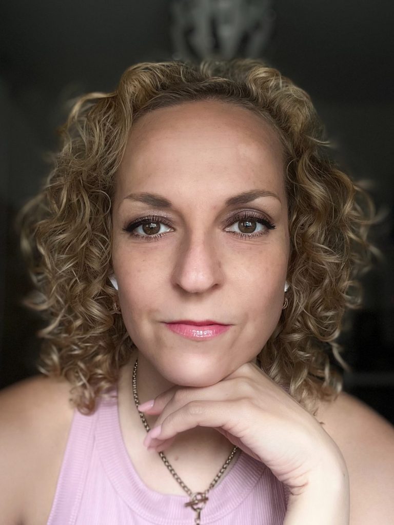 Porträt einer Frau mit lockigem, blonden Haar, neutralem Ausdruck und pinkem Lippenstift.